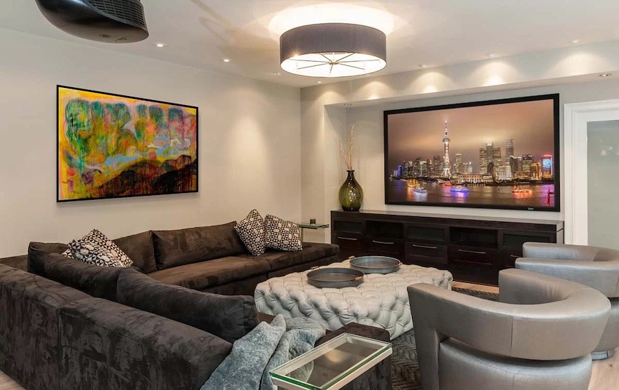 Media room with large television and artwork on the walls, sectional couch and two chairs, and large ceiling light fixture and recessed LED ceiling lighting.